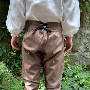 Pode incluir: Calções castanhos até ao joelho com cintura subida e fecho de atacadores pretos no centro das costas, combinados com uma camisa branca de manga comprida com punhos franzidos, usados sobre leggings pretas, com um design histórico.