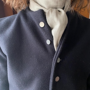 Puede incluir: Una chaqueta azul oscuro con cuello alto y botones plateados. Una bufanda beige claro está atada alrededor del cuello.