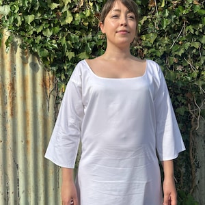 May include: A plain white dress featuring a wide scoop neckline and three-quarter length sleeves. The garment is constructed from a smooth, solid-coloured fabric that extends to a full-length hem, providing a simple and clean silhouette.