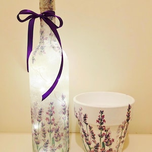 May include: A decorative glass bottle with a cork stopper and a purple ribbon, adorned with lavender sprigs and fairy lights. Beside it is a matching white flower pot, also decorated with lavender. The items are ideal for home decor.