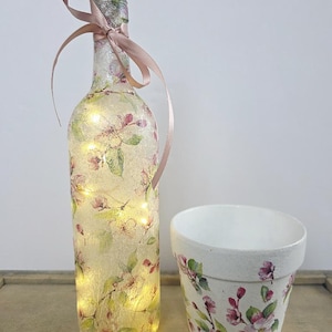 May include: A glass bottle with a textured finish, decorated with a pink and green floral pattern and illuminated by internal string lights, featuring a light pink ribbon tied around the neck, alongside a matching white ceramic flower pot.