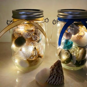 May include: Two glass mason jars filled with seashells, sand, and fairy lights. One jar has a gold ribbon, the other a blue ribbon. A large seashell rests in front of the jars, creating a coastal theme.