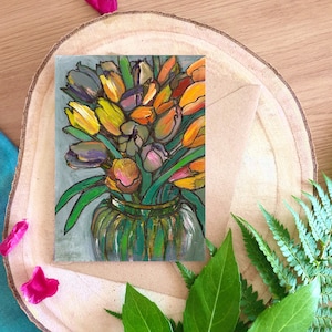 May include: A greeting card with a painting of tulips in a green vase. The tulips are yellow, orange, purple, and pink, with green leaves. The card sits on a wooden slice, with a kraft paper envelope alongside.