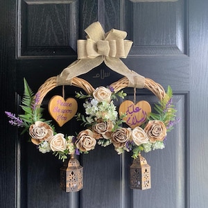 May include: A brown wicker heart-shaped wreath with faux flowers and greenery. Two wooden hearts hang from the wreath, one with the text "Peace Be Upon You" and the other with Arabic calligraphy. Two gold lanterns hang below the wreath.