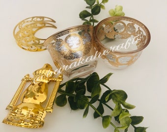 May include: A set of three gold-colored decorative items: a crescent moon-shaped tray, a small glass cup with gold and silver designs, and a gold-colored lantern-shaped ornament. The items are arranged on a white surface with green foliage.
