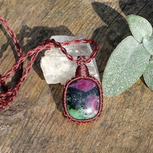 Puede incluir: Un colgante de piedra preciosa verde, rosa y negra en un collar de cordón marrón. El colgante tiene forma ovalada y está engastado en un nudo macramé.