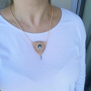Op de afbeelding: Een beige macramé ketting met een driehoekige hanger met een ronde, iriserende edelsteen. De hanger heeft een zilverkleurige punt aan de onderkant.