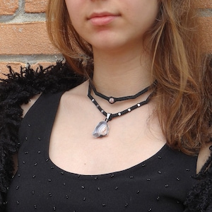 Può includere: Una donna indossa una camicetta nera con una collana a più strati con un ciondolo a forma di conchiglia e un accento di pietra rosa. La collana è realizzata con un cordoncino nero e una catena d'argento.