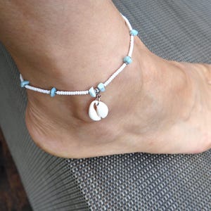 Può includere: Cavigliera di perline bianche con accenti azzurri e un ciondolo a forma di conchiglia. La cavigliera è indossata su un piede, mettendo in mostra il design delicato. Un accessorio a tema spiaggia.