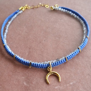 Può includere: Una collana di perline blu e trasparenti con un ciondolo a forma di mezzaluna d'oro.