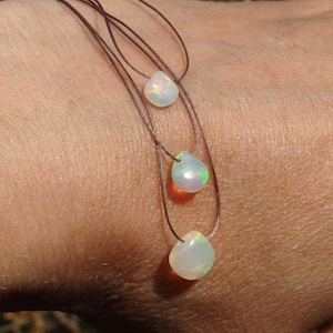 Könnte beinhalten: Drei zarte Halsketten, jede mit einem tropfenförmigen Opalanhänger. Die Opale zeigen ein Farbspiel mit grünen und orangenen Reflexen. Die Halsketten sind an dünnen, braunen Kordeln befestigt. Jeder Anhänger ist etwa 1 cm lang.