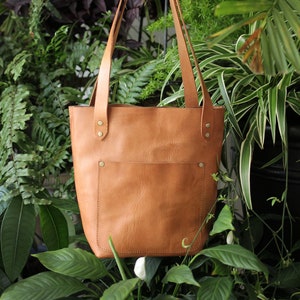 Puede incluir: Un bolso tote de cuero color marr&oacute;n claro con un bolsillo frontal y correas largas. El bolso se sostiene frente a un fondo de plantas verdes. El bolso tiene remaches de lat&oacute;n y una peque&ntilde;a letra "C" en relieve.