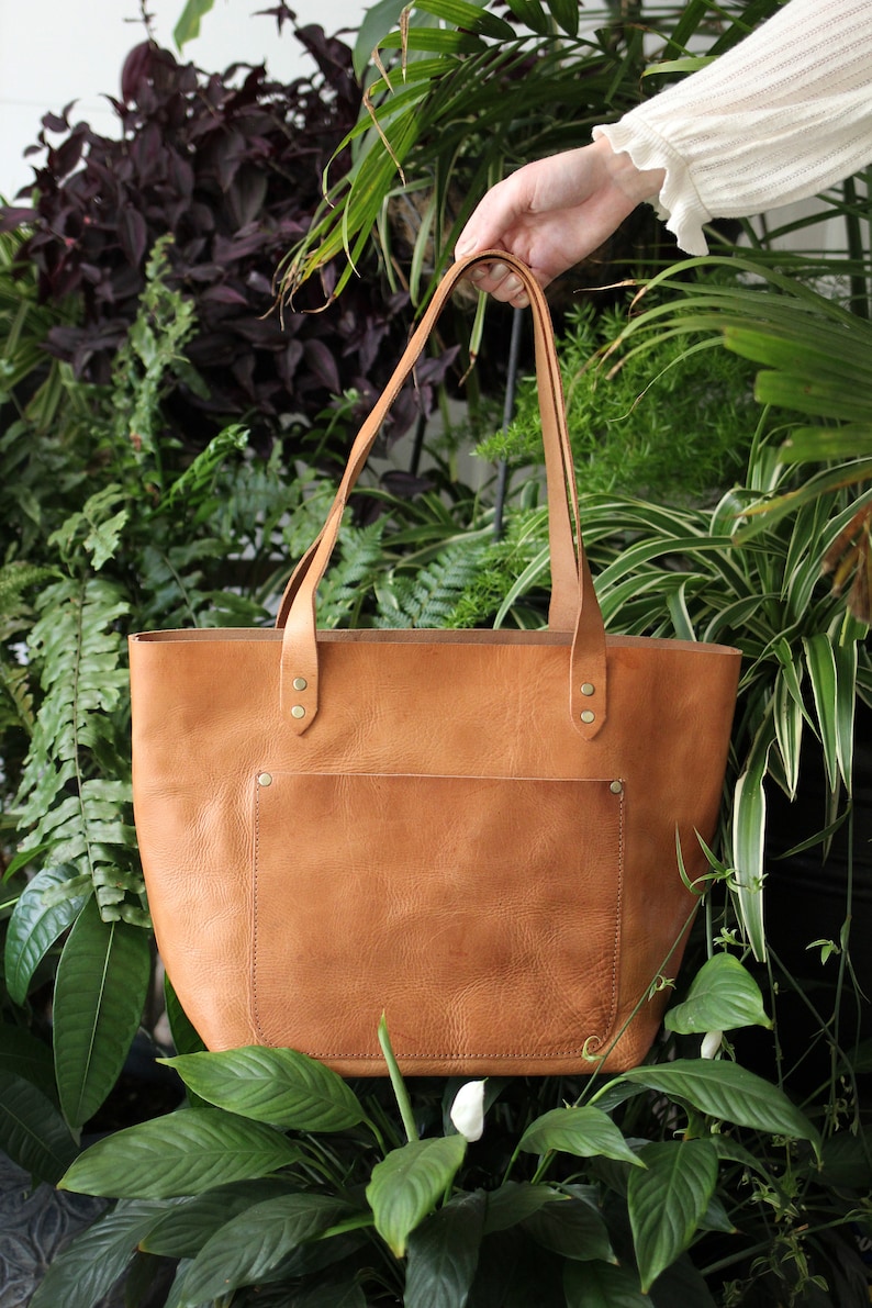 Puede incluir: Un bolso tote de cuero marr&oacute;n con un bolsillo frontal y herrajes dorados. El bolso est&aacute; sostenido por una mano sobre un fondo de plantas verdes.
