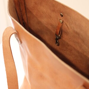 Puede incluir: Un bolso tote de cuero marr&oacute;n claro con una sola correa y un gancho de lat&oacute;n en el interior. El bolso est&aacute; abierto y el interior es visible.