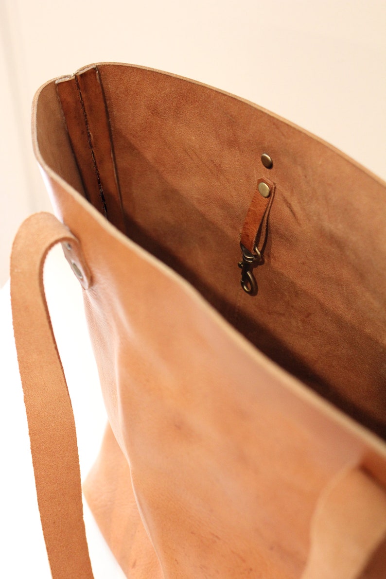 Puede incluir: Un bolso tote de cuero marr&oacute;n claro con una sola correa y un gancho de lat&oacute;n en el interior. El bolso est&aacute; abierto y el interior es visible.