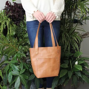 Puede incluir: Un bolso tote de cuero color canela con un bolsillo frontal, sostenido por una persona. El bolso tiene correas largas y est&aacute; asegurado con remaches. La persona lleva vaqueros azules y botas color canela. El fondo presenta varias plantas verdes.
