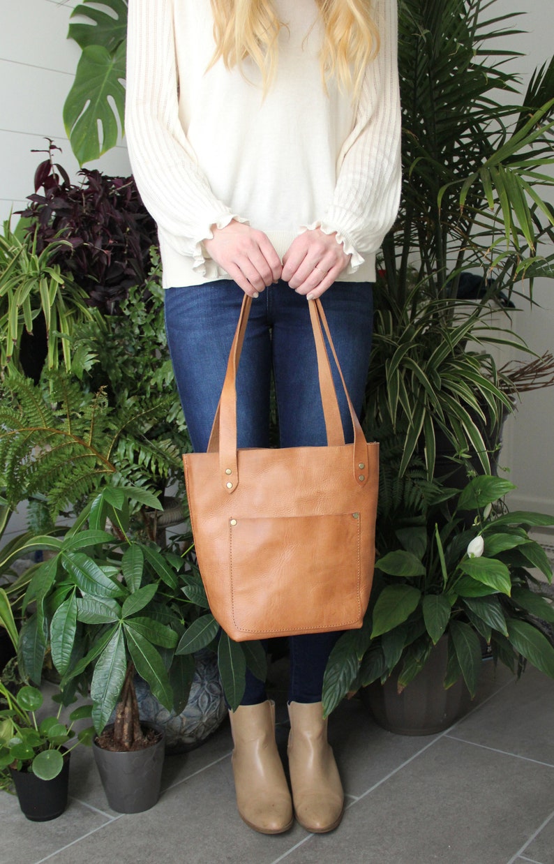 Puede incluir: Un bolso tote de cuero color canela con un bolsillo frontal, sostenido por una persona. El bolso tiene correas largas y est&aacute; asegurado con remaches. La persona lleva vaqueros azules y botas color canela. El fondo presenta varias plantas verdes.