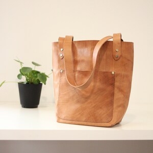 Puede incluir: Un bolso tote de cuero marr&oacute;n claro con un bolsillo frontal y dos asas. El bolso tiene forma rectangular y est&aacute; hecho de cuero texturizado. Las asas est&aacute;n sujetas con remaches. Una peque&ntilde;a planta en maceta est&aacute; en el fondo.