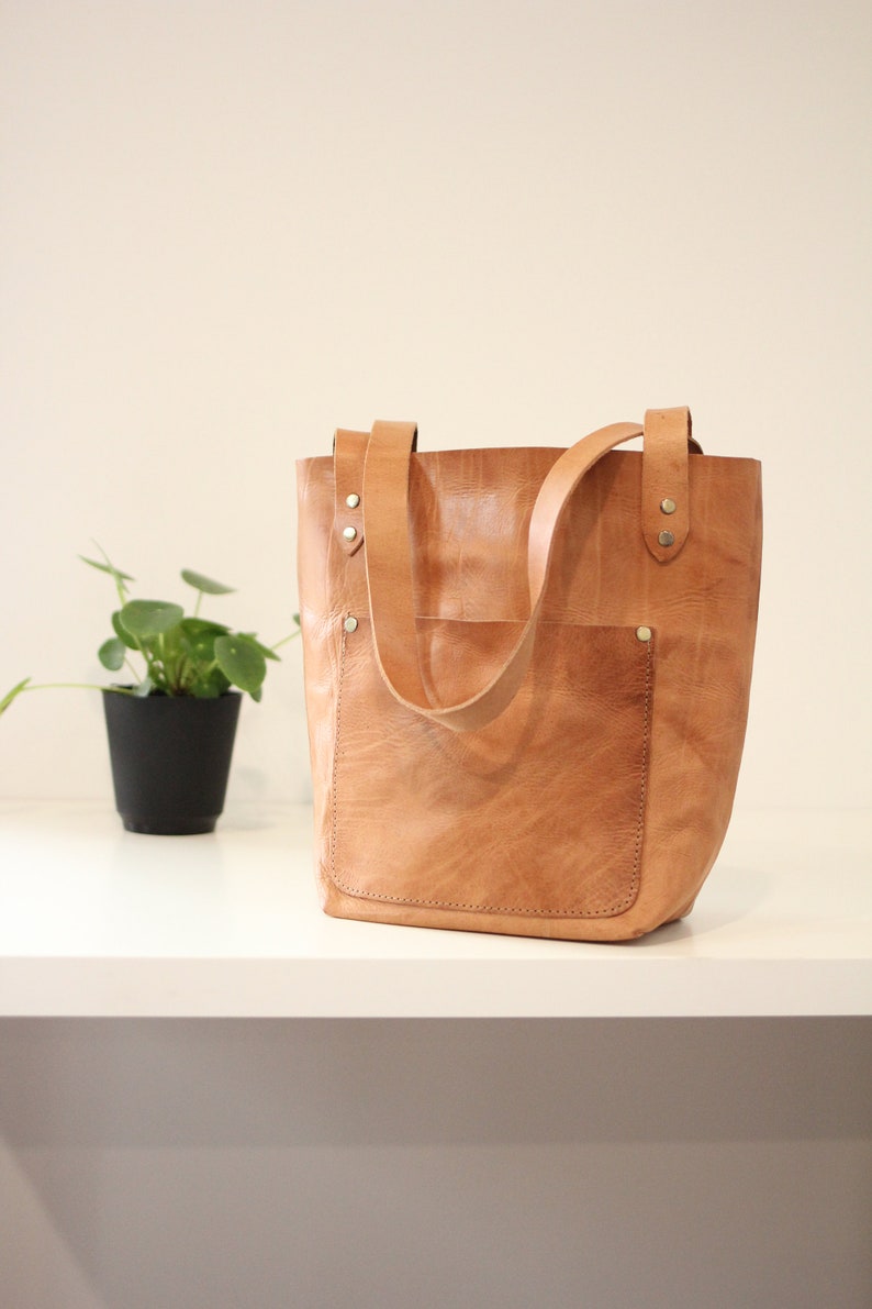 Puede incluir: Un bolso tote de cuero marr&oacute;n claro con un bolsillo frontal y dos asas. El bolso tiene forma rectangular y est&aacute; hecho de cuero texturizado. Las asas est&aacute;n sujetas con remaches. Una peque&ntilde;a planta en maceta est&aacute; en el fondo.