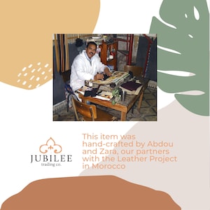 Puede incluir: Un hombre con bata blanca trabajando en una mesa de madera. El texto dice: "Este art&iacute;culo fue hecho a mano por Abdou y Zara, nuestros socios con el Leather Project en Marruecos". Tambi&eacute;n es visible el logotipo de Jubilee Trading Co.
