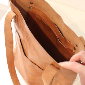 Puede incluir: Un bolso tote de cuero marr&oacute;n claro con dos correas largas y bolsillos interiores. El bolso est&aacute; abierto, revelando el interior y los bolsillos. El cuero tiene un aspecto natural y texturizado.
