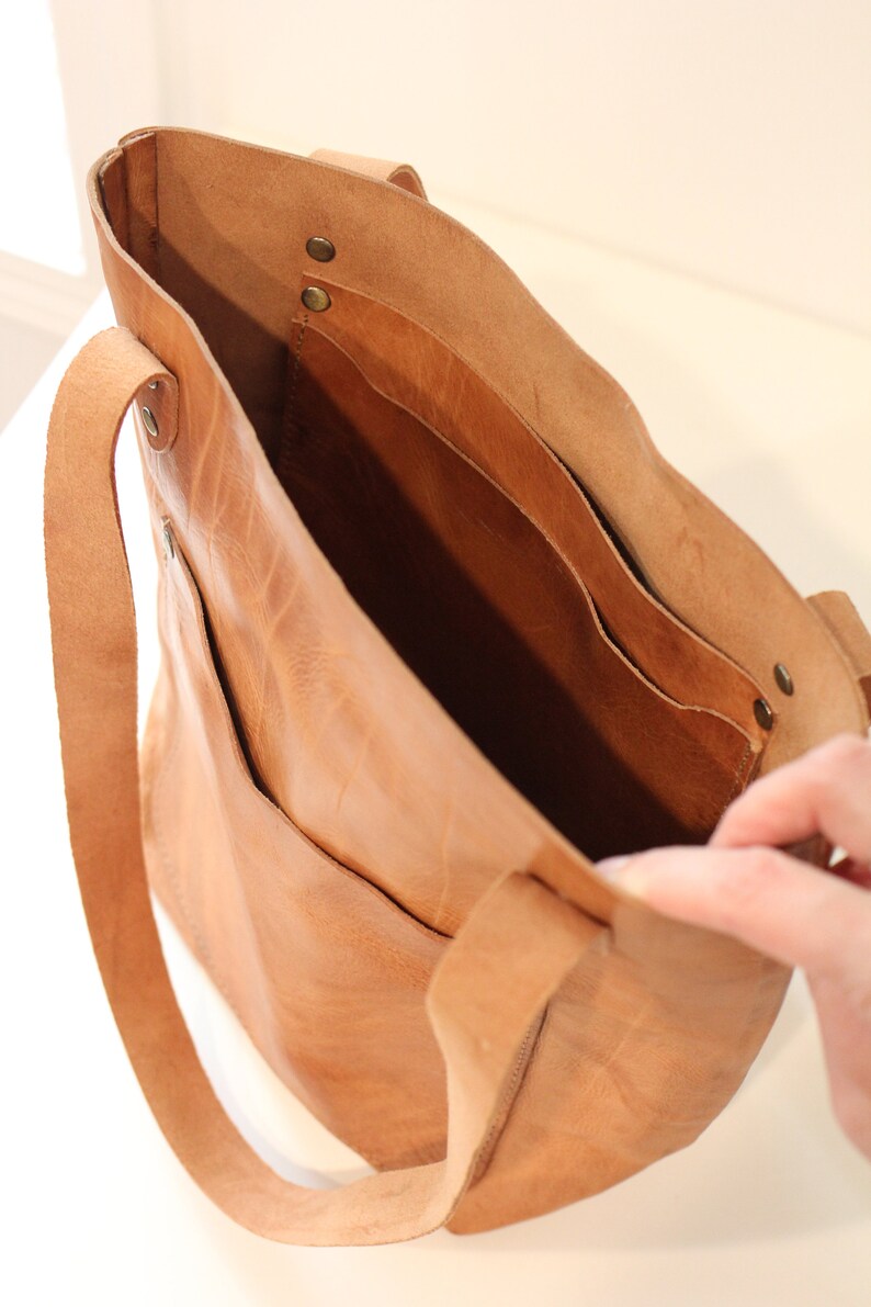 Puede incluir: Un bolso tote de cuero marr&oacute;n claro con dos correas largas y bolsillos interiores. El bolso est&aacute; abierto, revelando el interior y los bolsillos. El cuero tiene un aspecto natural y texturizado.