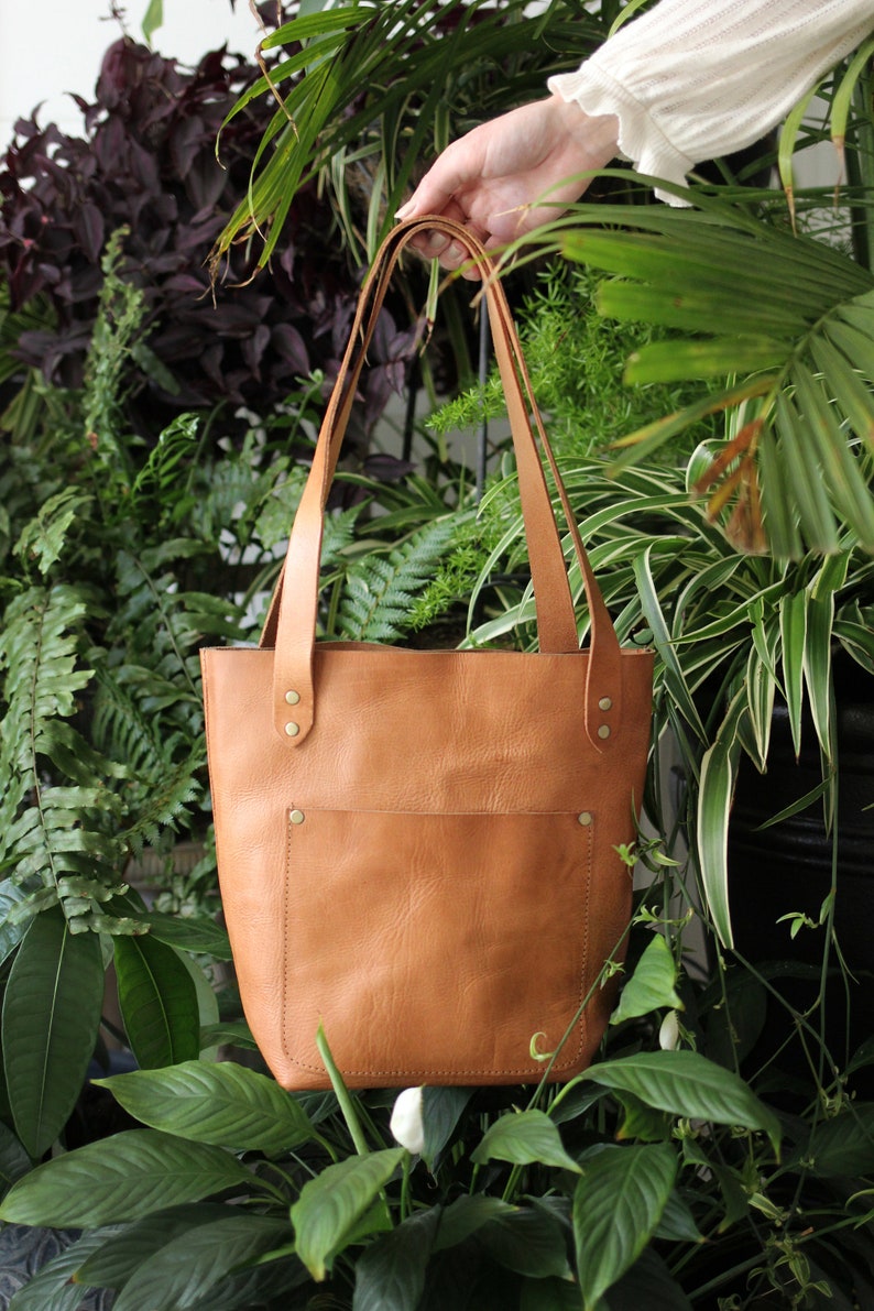 Puede incluir: Un bolso tote de cuero color marr&oacute;n claro con un bolsillo frontal y correas largas. El bolso se sostiene frente a un fondo de plantas verdes. El bolso tiene remaches de lat&oacute;n y una peque&ntilde;a letra "C" en relieve.