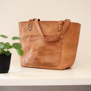 Puede incluir: Un bolso tote de cuero marr&oacute;n con un bolsillo frontal grande y dos asas superiores. El bolso tiene un aspecto desgastado y est&aacute; hecho de cuero de alta calidad.