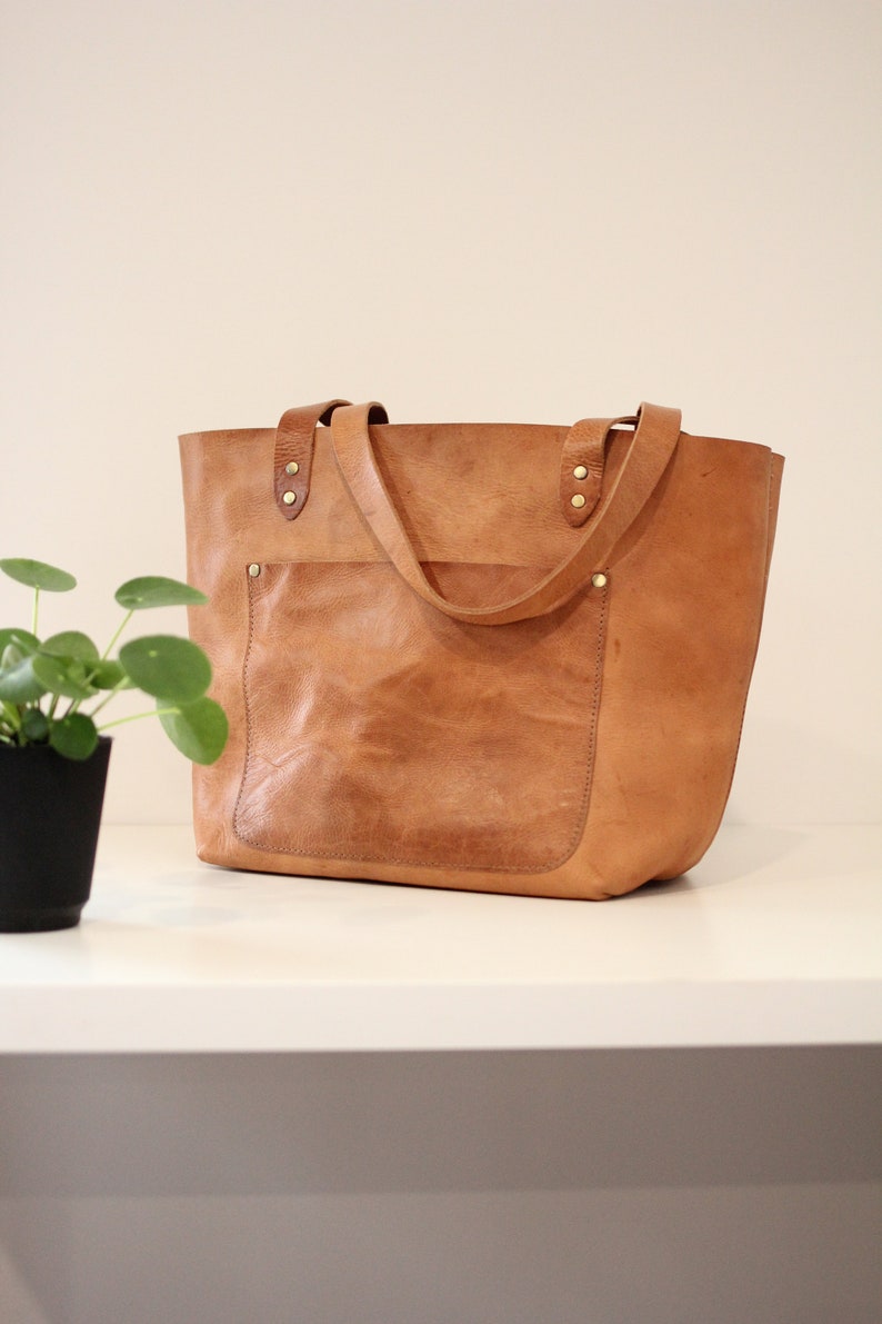 Puede incluir: Un bolso tote de cuero marr&oacute;n con un bolsillo frontal grande y dos asas superiores. El bolso tiene un aspecto desgastado y est&aacute; hecho de cuero de alta calidad.