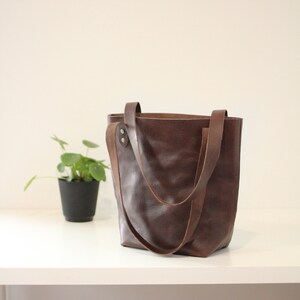 Puede incluir: Un bolso tote de cuero marr&oacute;n con dos asas. El bolso est&aacute; hecho de cuero texturizado y tiene un dise&ntilde;o simple y cl&aacute;sico. El bolso est&aacute; sobre una superficie blanca. Una peque&ntilde;a planta en maceta est&aacute; en el fondo.