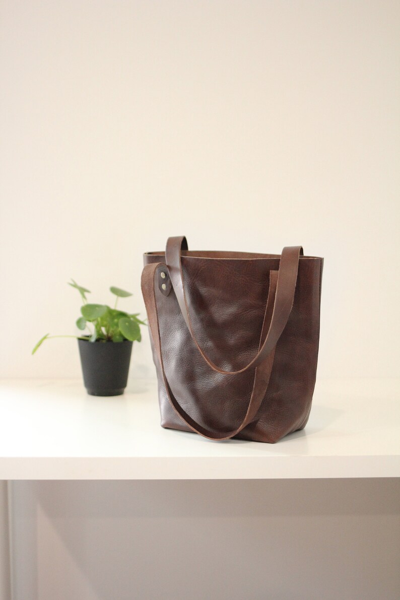 Puede incluir: Un bolso tote de cuero marr&oacute;n con dos asas. El bolso est&aacute; hecho de cuero texturizado y tiene un dise&ntilde;o simple y cl&aacute;sico. El bolso est&aacute; sobre una superficie blanca. Una peque&ntilde;a planta en maceta est&aacute; en el fondo.