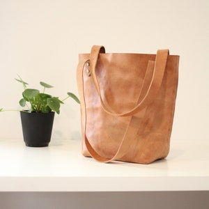 Puede incluir: Un bolso tote de cuero marr&oacute;n claro con correas largas. La bolsa est&aacute; sobre una superficie blanca junto a una peque&ntilde;a planta en maceta con hojas verdes. La bolsa tiene una apariencia texturizada.