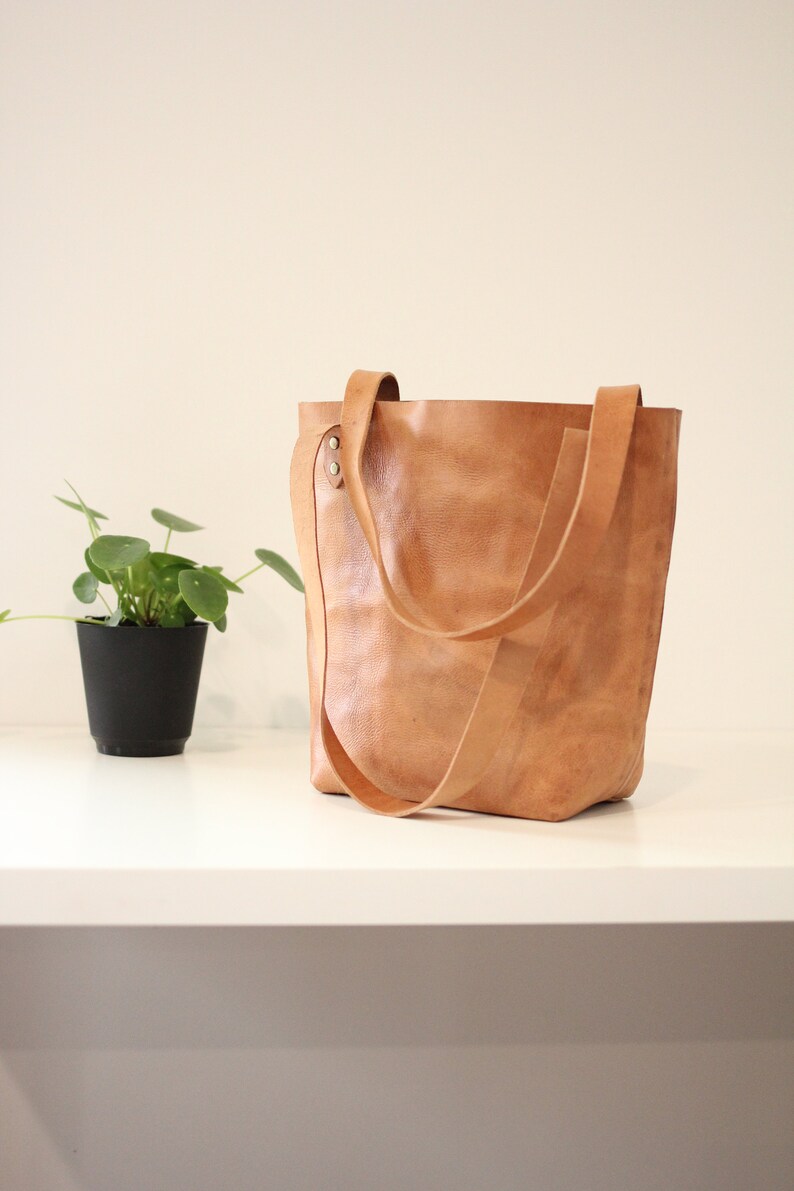 Puede incluir: Un bolso tote de cuero marr&oacute;n claro con correas largas. La bolsa est&aacute; sobre una superficie blanca junto a una peque&ntilde;a planta en maceta con hojas verdes. La bolsa tiene una apariencia texturizada.