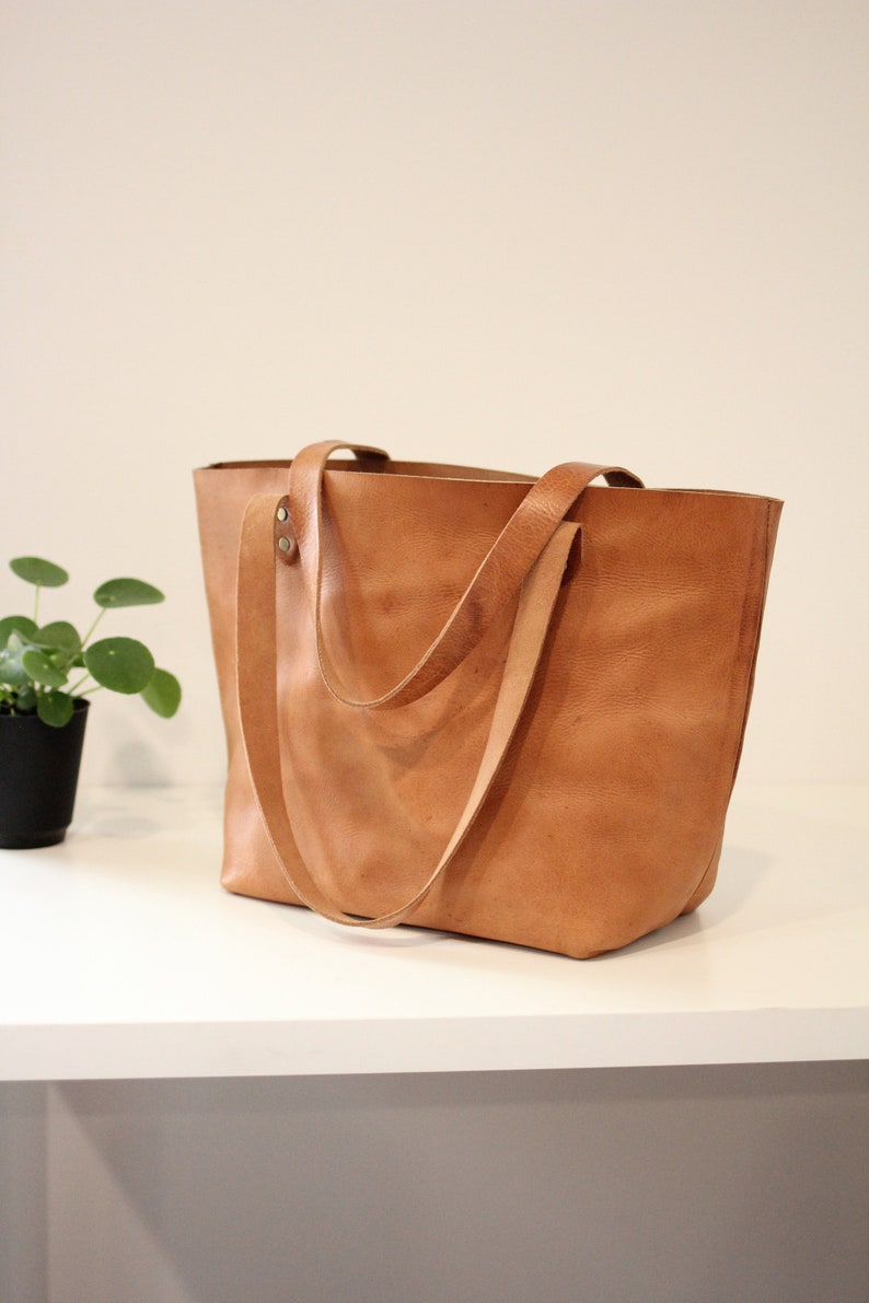 Puede incluir: Un bolso tote de cuero marr&oacute;n claro con dos asas largas. El bolso est&aacute; sobre una superficie blanca.