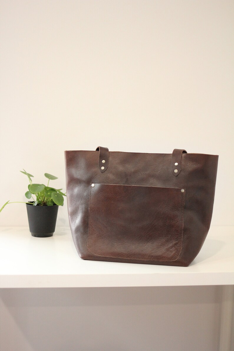 Puede incluir: Un bolso tote de cuero marr&oacute;n con un bolsillo frontal grande y remaches plateados. El bolso tiene dos asas y est&aacute; sobre una estanter&iacute;a blanca.