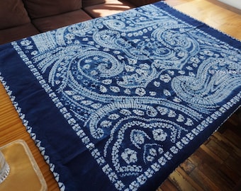 Könnte beinhalten: Ein quadratisches, indigoblaues Textil mit einem weißen, paisleyartigen Muster. Der Stoff liegt auf einem Holztisch mit einem dunkelblauen Rand. Das Design zeigt wirbelnde Formen und florale Motive und schafft eine dekorative Tischdecke.