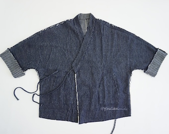 Könnte beinhalten: Dunkelblaue Jacke im Kimono-Stil mit strukturiertem Gewebe. Die Ärmel sind umgeschlagen und zeigen ein gemustertes Futter. Wickeldesign mit Bindebändern und einer kleinen Tasche. Der Text "@YoulanStudio" ist unten rechts sichtbar.