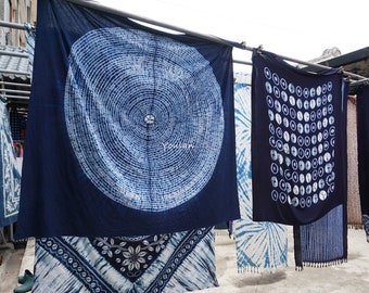 Könnte beinhalten: Im Freien hängen indigogefärbte Textilien. Die Stoffe zeigen verschiedene weiße Muster auf einem dunkelblauen Hintergrund, darunter konzentrische Kreise, geometrische Formen und florale Designs. Das Wort "Youlan" ist auf einem der Stoffe zu sehen.