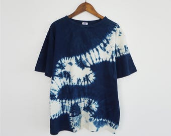 Könnte beinhalten: Ein kurzärmeliges T-Shirt mit einem dunkel marineblauen und weißen Batikmuster, das ein diagonales, geschwungenes Design auf dem Stoff erzeugt. Das T-Shirt hat einen Rundhalsausschnitt und wird auf einem Holzkleiderbügel vor weißem Hintergrund präsentiert.