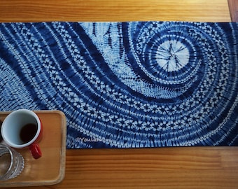 Könnte beinhalten: Blau-weißes Tischläufer mit Batikmuster und Spiraldesign. Der Läufer liegt auf einem Holztisch, mit einem Holztablett, einer Tasse mit rotem Griff und einem Glasbehälter im Vordergrund.