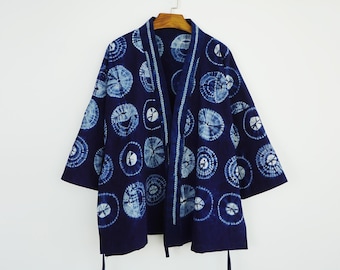 Könnte beinhalten: Marineblaue Kimono-Jacke mit einem Batikmuster aus weißen und hellblauen Kreisen. Die Jacke hat eine lockere Passform mit weiten Ärmeln und einem kragenlosen Design. Sie hängt an einem Holzbügel vor weißem Hintergrund.