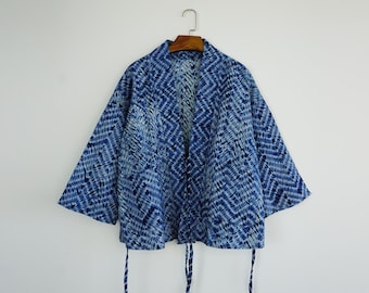 Könnte beinhalten: Blau-weiß gemusterte Kimono-Jacke mit weiten Ärmeln, an einem Holzbügel hängend. Die Jacke hat einen Bindeverschluss vorne und ein geometrisches Design. Der Stoff scheint leicht zu sein, ideal zum Überziehen.