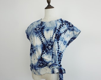 Könnte beinhalten: Ein weißes und indigoblaues Batik-Top. Das kurzärmelige Shirt hat ein Wickeldesign mit Bindeband in der Taille. Der Stoff ist mit kreisförmigen Mustern versehen, was einen künstlerischen Look erzeugt. Das Top wird auf einer Schaufensterpuppe präsentiert.