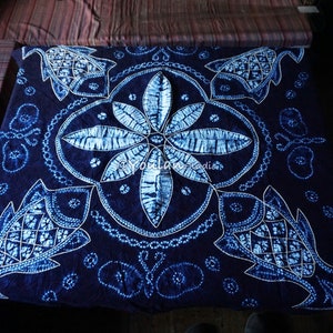 Puede incluir: Una tela teñida en índigo azul y blanco con un diseño japonés tradicional. La tela presenta un motivo floral central rodeado de cuatro diseños de peces. La tela es de forma cuadrada y tiene un borde con flecos.