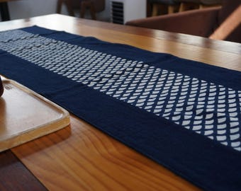 Könnte beinhalten: Ein langer, rechteckiger Tischläufer aus dunkelblauem Stoff mit einem Mittelstreifen, der mit einem sich wiederholenden weißen geometrischen Muster aus kleinen, gestapelten, geschwungenen Formen verziert ist, die an stilisierte Fischschuppen erinnern.