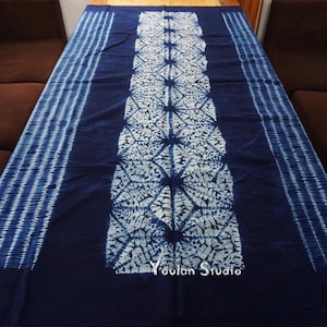 Op de afbeelding: Een donkerblauwe textiel met een wit patroon. Het centrale gedeelte heeft een herhalend geometrisch patroon, terwijl de randen verticale strepen hebben. De textiel wordt gepresenteerd op een houten oppervlak, met de woorden "Youlan Studio" zichtbaar.
