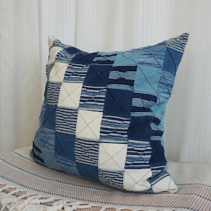 Op de afbeelding: Vierkant decoratief kussen met een patchwork ontwerp in blauw- en wittinten. Het kussen heeft een verscheidenheid aan gestructureerde stoffen en patronen, waaronder strepen en vierkanten. Het kussen ligt op een textiel met franjes.