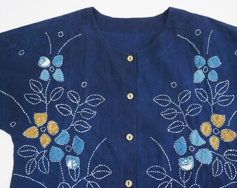Könnte beinhalten: Ein marineblaues Hemd mit Knöpfen und kurzen Ärmeln. Das Hemd hat weiße, gestickte Blumendesigns mit blauen und goldenen Akzenten. Das Design ist auf beiden Seiten des Hemdes symmetrisch.