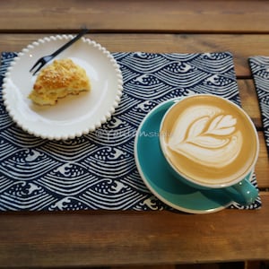 Könnte beinhalten: Ein weißer Teller mit einem Scone auf einem blauen und weißen Platzdeckchen mit Wellenmuster. Eine Tasse Kaffee mit Latte Art steht auf einem türkisfarbenen Untersetzer auf einem ähnlichen Platzdeckchen.
