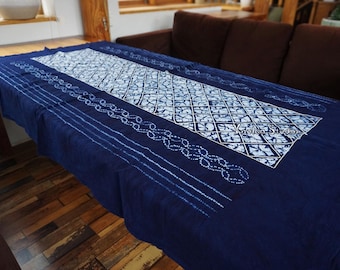 Könnte beinhalten: Eine marineblaue Tischdecke mit einem weißen geometrischen Muster in der Mitte. Die Ränder weisen eine Reihe weißer gepunkteter Linien und ein kettenartiges Design auf. Die Tischdecke ist über einem Holztisch drapiert.